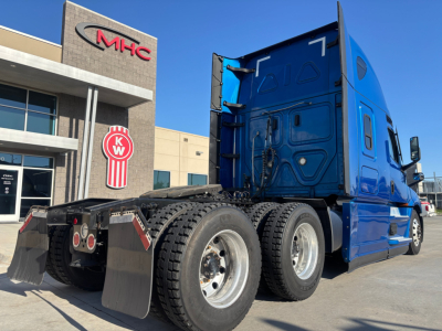 Freightliner PT126064ST tractor rear rear-axle view showing dual rear wheels and mudflaps; blue sleeper cab with stacked exhaust and trailer hitch, dealership building in background.