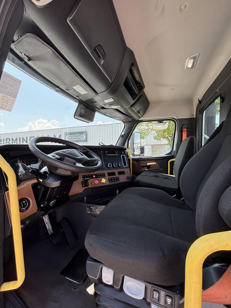 Cab interior of a 2023 Freightliner Cascadia 126 truck showing steering wheel, dashboard with controls, driver seat, and passenger seat; beige and black color scheme; dashboard gauges and overhead compartments visible.
