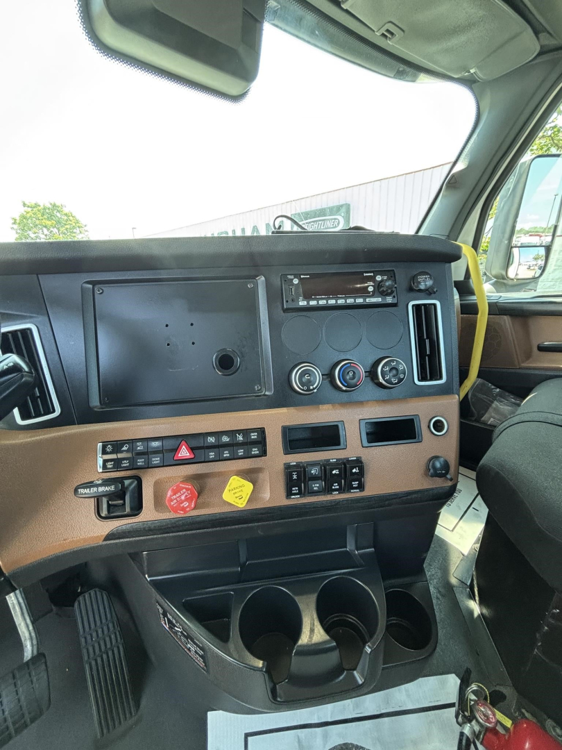 Cab interior of a Freightliner Cascadia showing dashboard with vents, gauges, cup holders, and control panel. Visible labels include hazard button and cup holders.