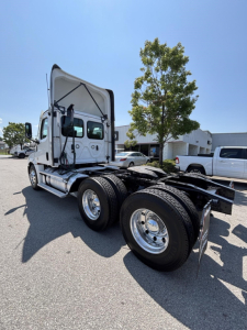 Freightliner Cascadia 126 day cab tractor rear three-quarter view with raised cab, dual rear axles, chrome wheels, and fuel tanks visible.
