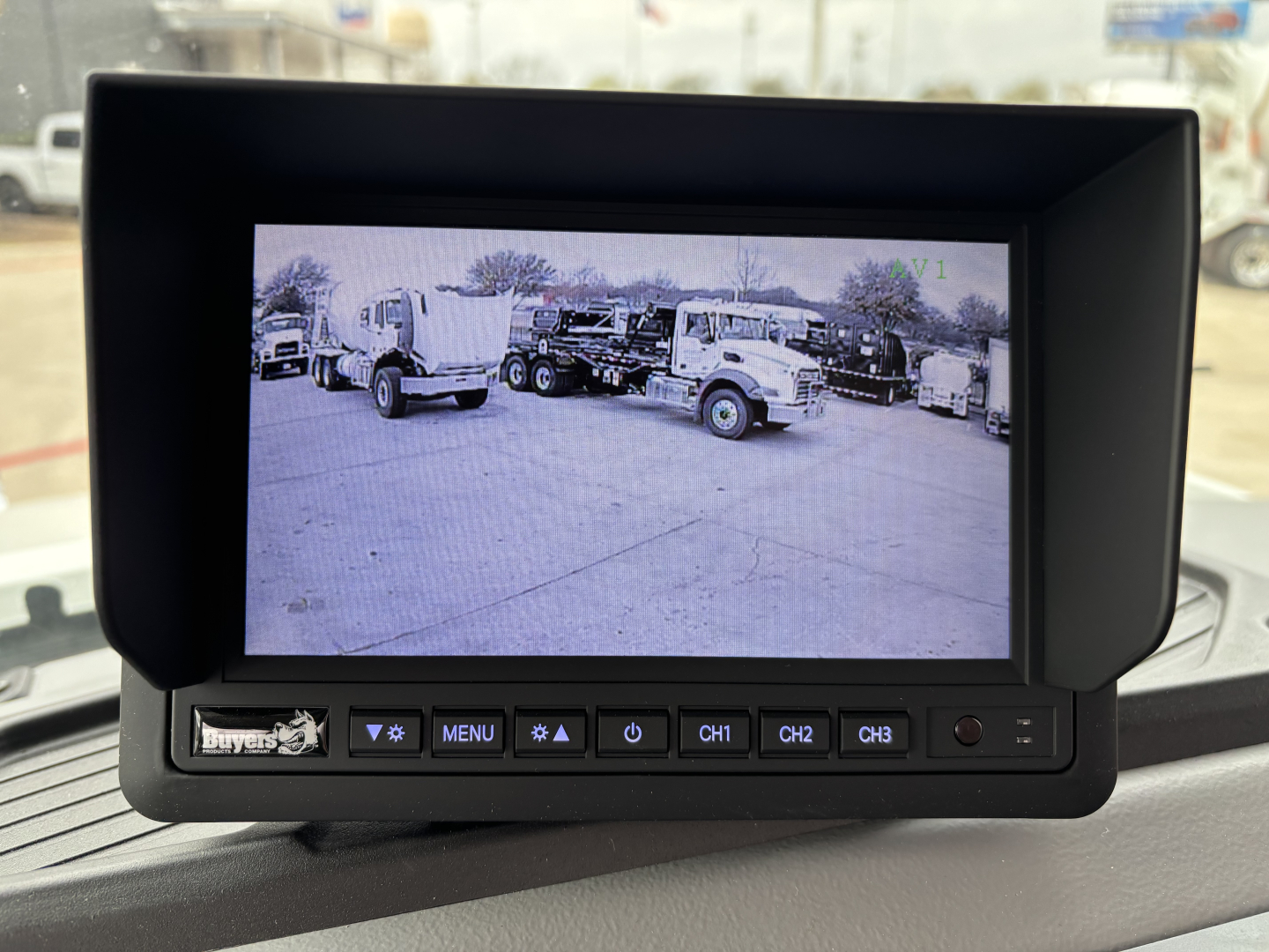 In-vehicle color display monitor with control buttons in front; screen shows a parking lot with trucks. Visible text on unit includes 'Bus' or 'Buyer' label on lower left and buttons labeled MENU, CH1, CH2, CH3. No other legible text clearl
