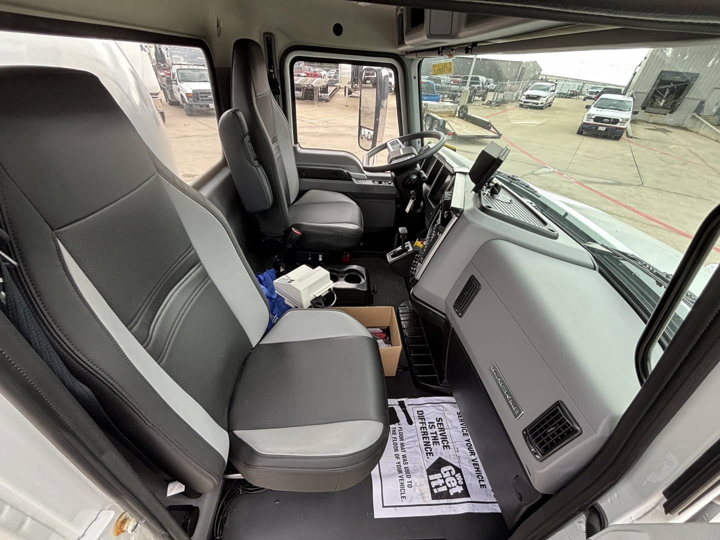 Truck cab interior of a 2026 Mack MD7 showing driver seat and passenger seat, steering wheel, dashboard, and center console with papers on floor mat.