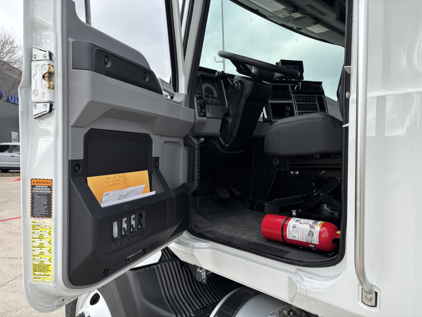 Cab interior of a 2026 Mack MD7 truck showing open door with storage pocket and steering area; fire extinguisher on passenger footwell and dashboard details visible.