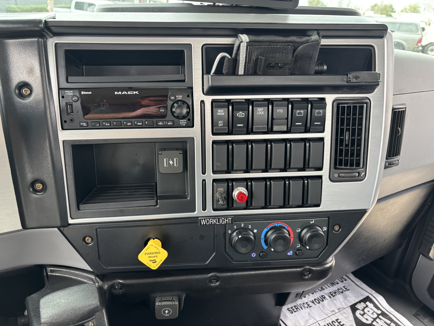 Dashboard panel of a 2026 Mack MD7 truck showing radio, switches, climate controls, and a yellow hazard sticker.
