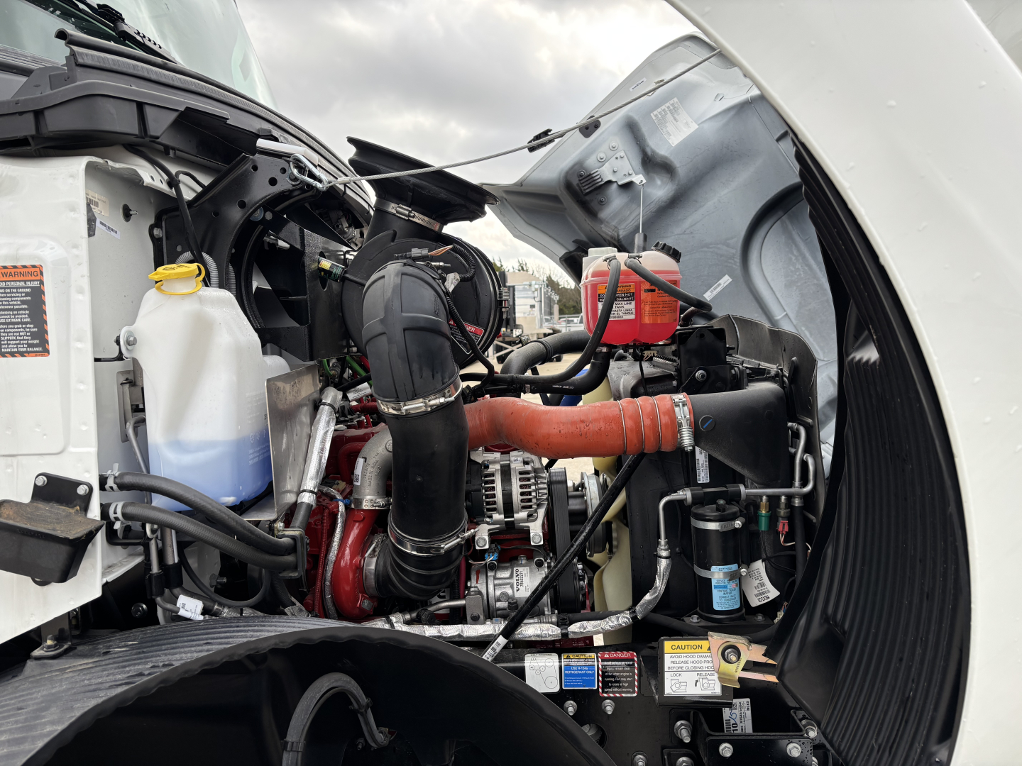 Engine bay of a new Mack MD7 truck showing coolant reservoir, black intake hose, orange intercooler pipe, alternator, and various labels/stickers.