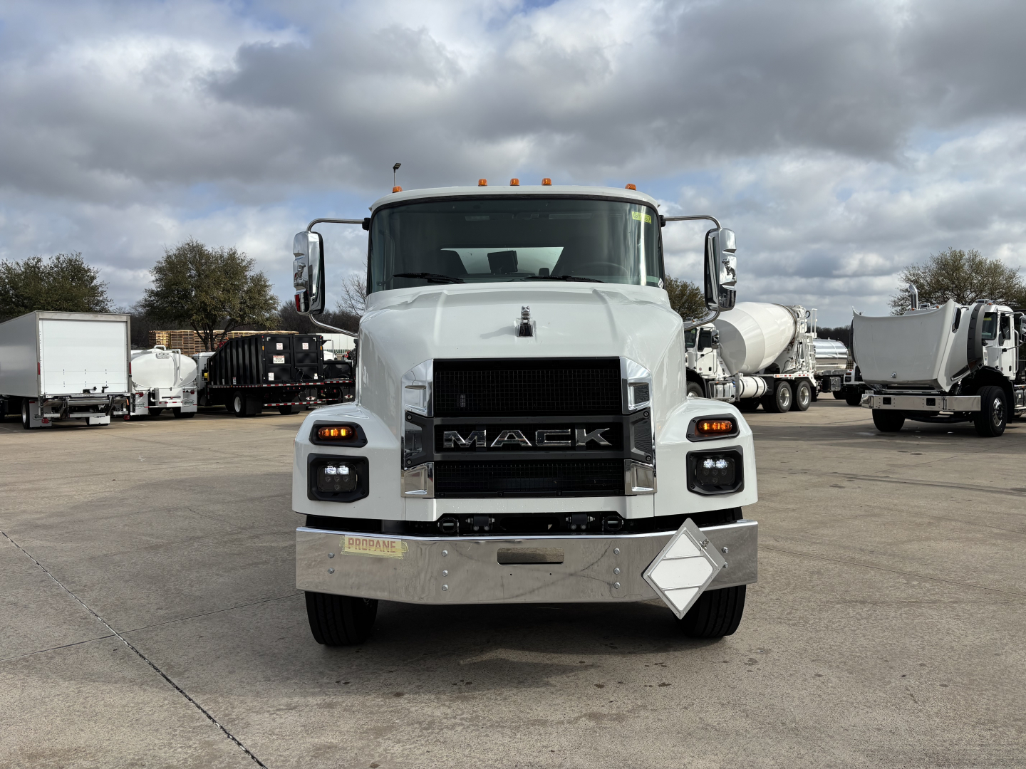 Front view of 2026 Mack MD7 truck in a lot, with large grille, headlights, chrome bumper, and diamond-shaped hazard plate on bumper.