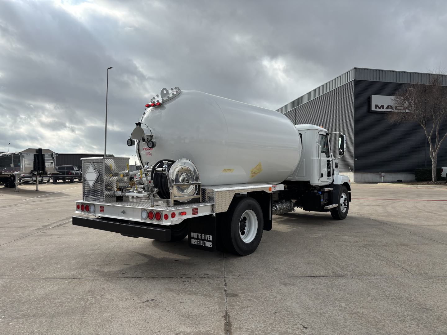 Tank truck with cylindrical rear tank, white cab, rear view showing rear bumper, taillights, and hoses. Mack MD7 dealership building in background.