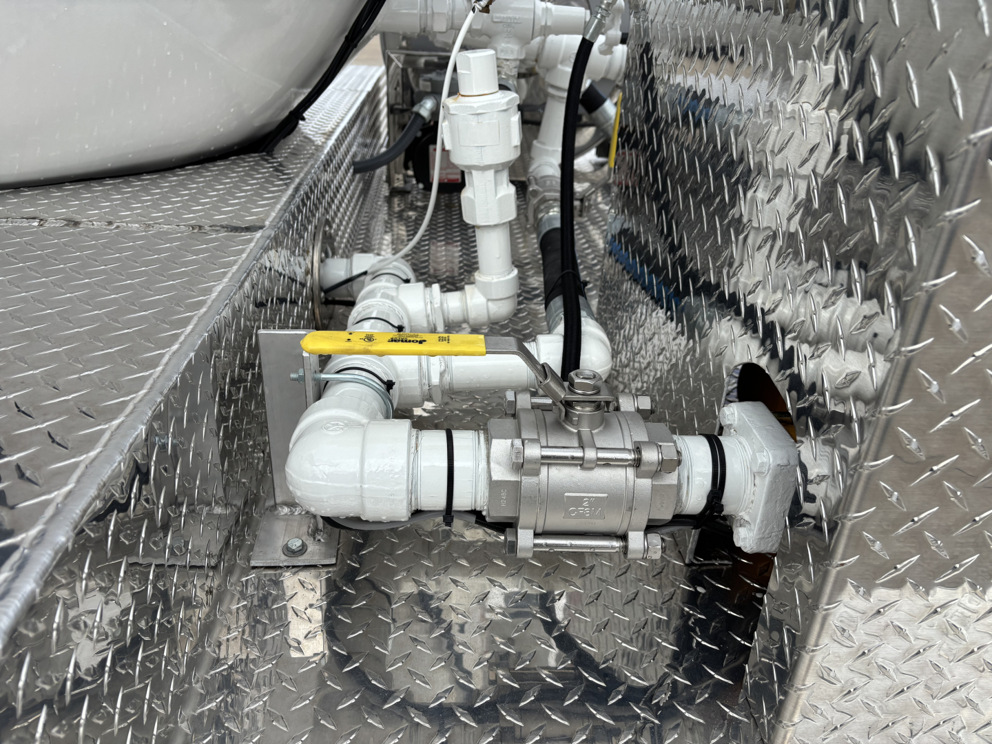 Machinery valve assembly and piping in a diamond-plate engine bay with a yellow-handled valve and inline fittings.