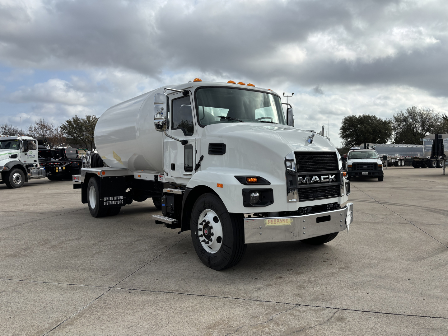 Mack MD7 concrete mixer truck in a dealership lot, front three-quarter view, white cab with black Mack grille, chrome bumper, orange clearance lights, background other trucks.