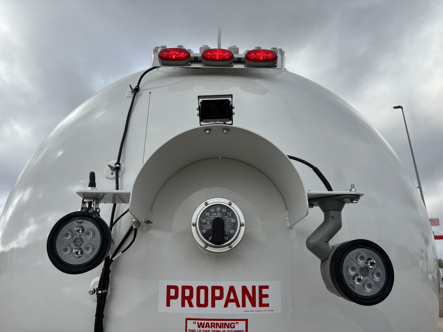Rear view of a white propane tank trailer with red lights on top, circular manway, and two circular LED/spotlights on sides. A large PROPANE label is centered below the manway.