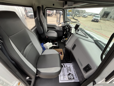 Truck cab interior of a 2026 Mack MD7 showing driver seat and passenger seat, steering wheel, dashboard, and center console with papers on floor mat.