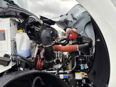 Engine bay of a new Mack MD7 truck showing coolant reservoir, black intake hose, orange intercooler pipe, alternator, and various labels/stickers.