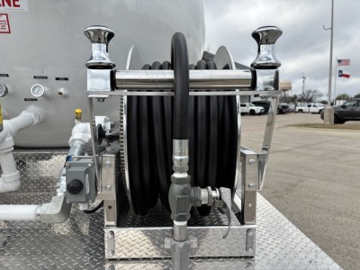 Front view of a Mack MD7 truck tank with a hose reel and chrome fittings; visible components include a black hose wound on a reel, metal brackets, fittings, and a diamond plate surface.
