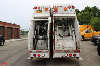 Back view of two Mack TerraPro cabover trucks, rear compartments open with equipment and hoses visible. License plates and taillights present. Dirt and grime on bodywork.