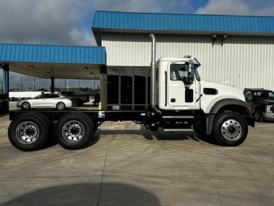 Truck side view of a 2027 Mack GR64F dump truck showing white cab and six-wheeled chassis with black frame, housed under a blue-roofed building.