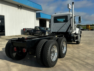 Mack GR64F dump truck chassis rear view showing triple axle and rear tires, metal frame, cab with exhaust stack, and parking lot.