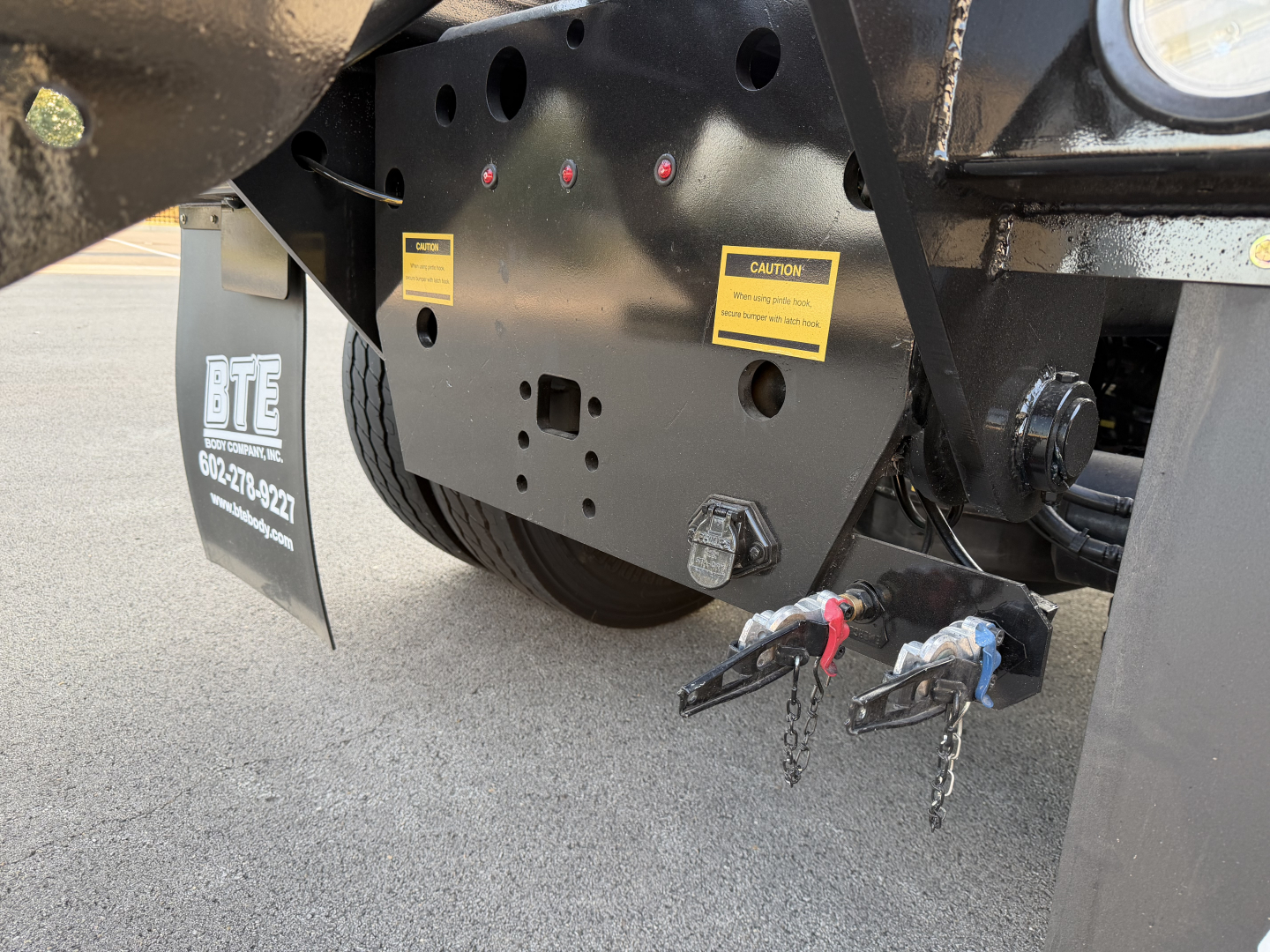 Mack GR64B rear view showing hitch pins and warning labels on the frame, black undercarriage with yellow caution stickers.