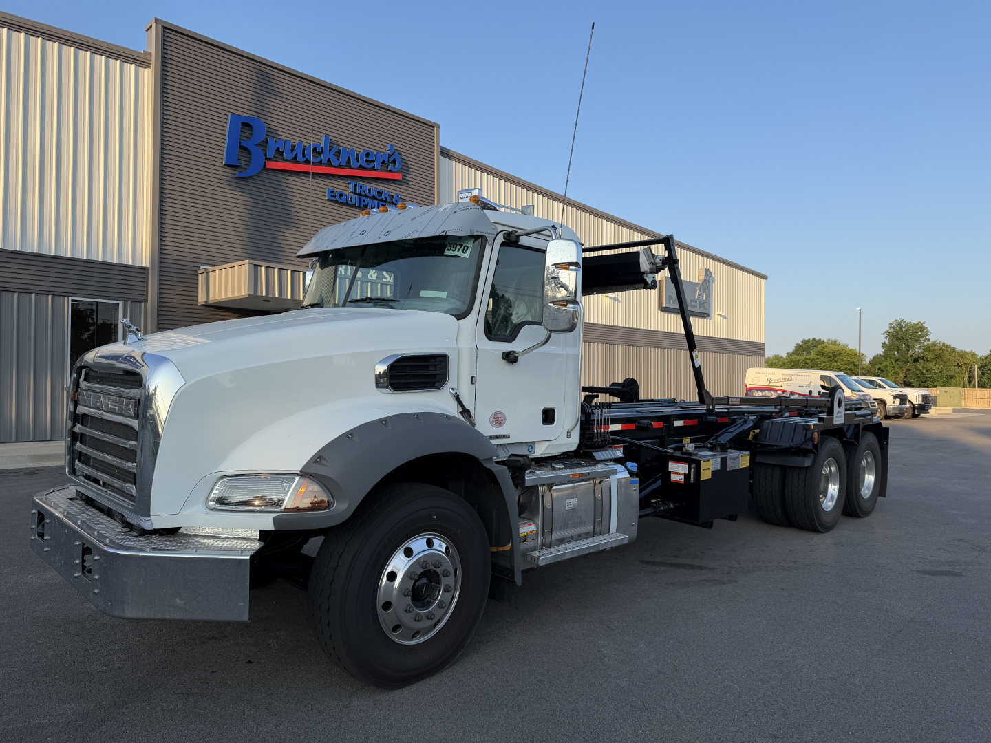 Truck: Mack GR64B rolloff installation showing white cab, black hooklift, and long chassis with dual rear axles parked in front of a dealership.