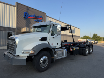 Truck: Mack GR64B rolloff installation showing white cab, black hooklift, and long chassis with dual rear axles parked in front of a dealership.