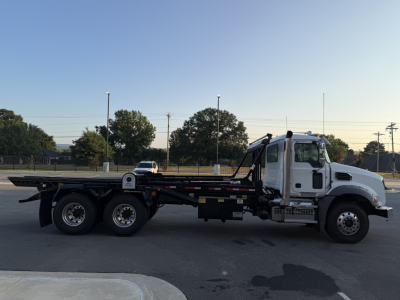 Mack GR64B rolloff truck in side profile showing long flatbed with dual rear axles and hydraulic mixer arm on the cab side.