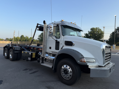 Mack GR64B roll-off truck in daylight showing side view with cab, chassis, and loading crane. Clear windshield, chrome exhaust stack, and dual rear axles.