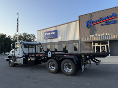 Mack GR64B rolloff truck in front of a dealership with Volvo Trucks sign, showing a long flatbed with multiple axles and dual rear wheels.