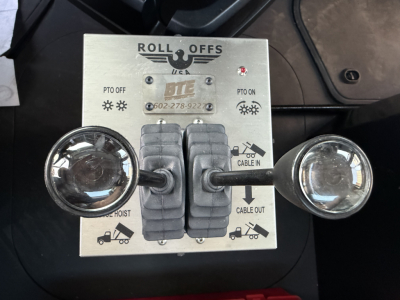 Roll Offs USA control panel with two circular lights, central dual levers, and indicator icons. Text includes ROLL OFFS USA, PTO OFF, PTO ON, CABLE IN, CABLE OUT, and symbols for hoist and trailer.