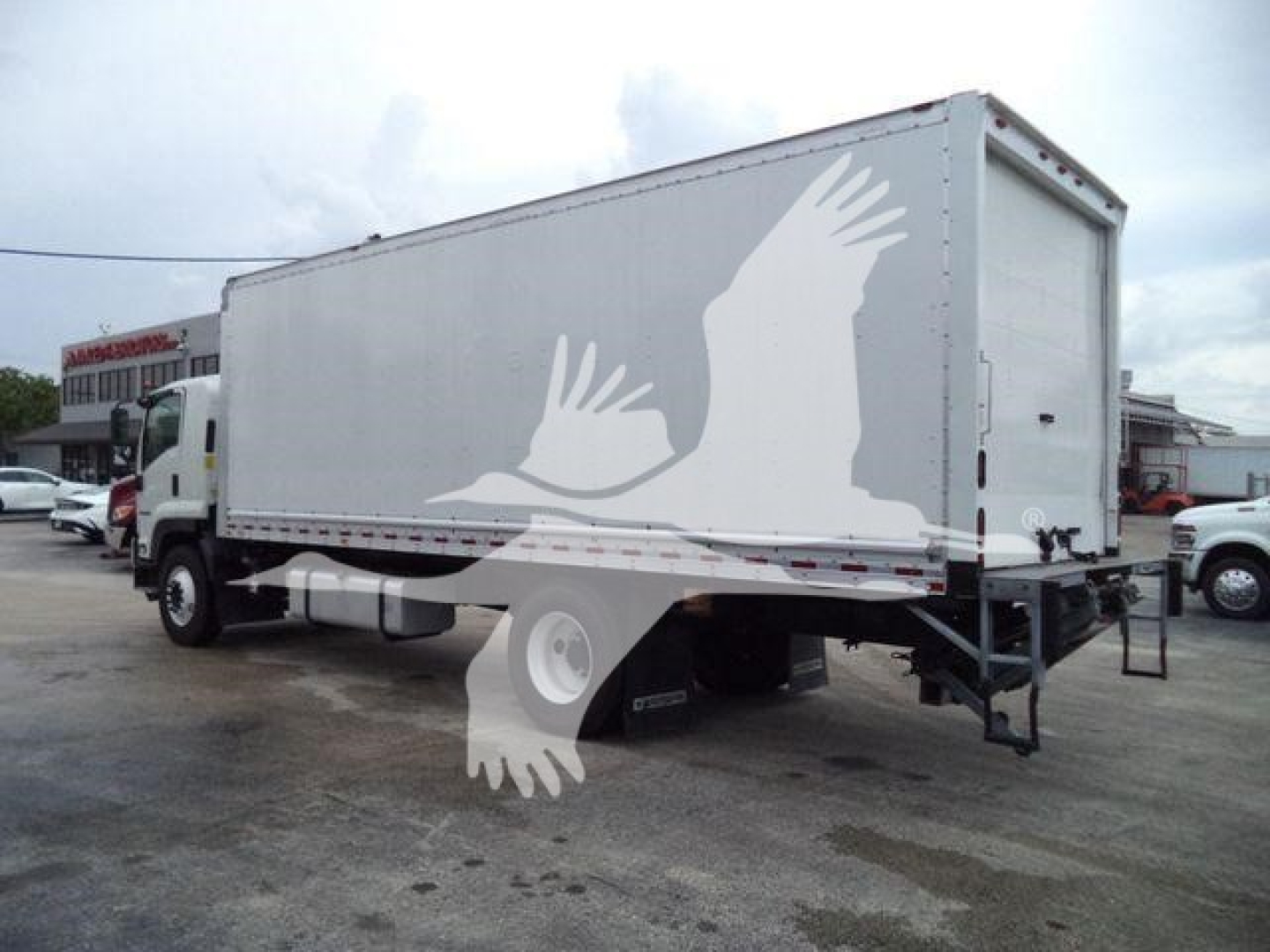Isuzu NPR or box truck with a large white cargo box, rear and side views, some traffic and buildings in background.