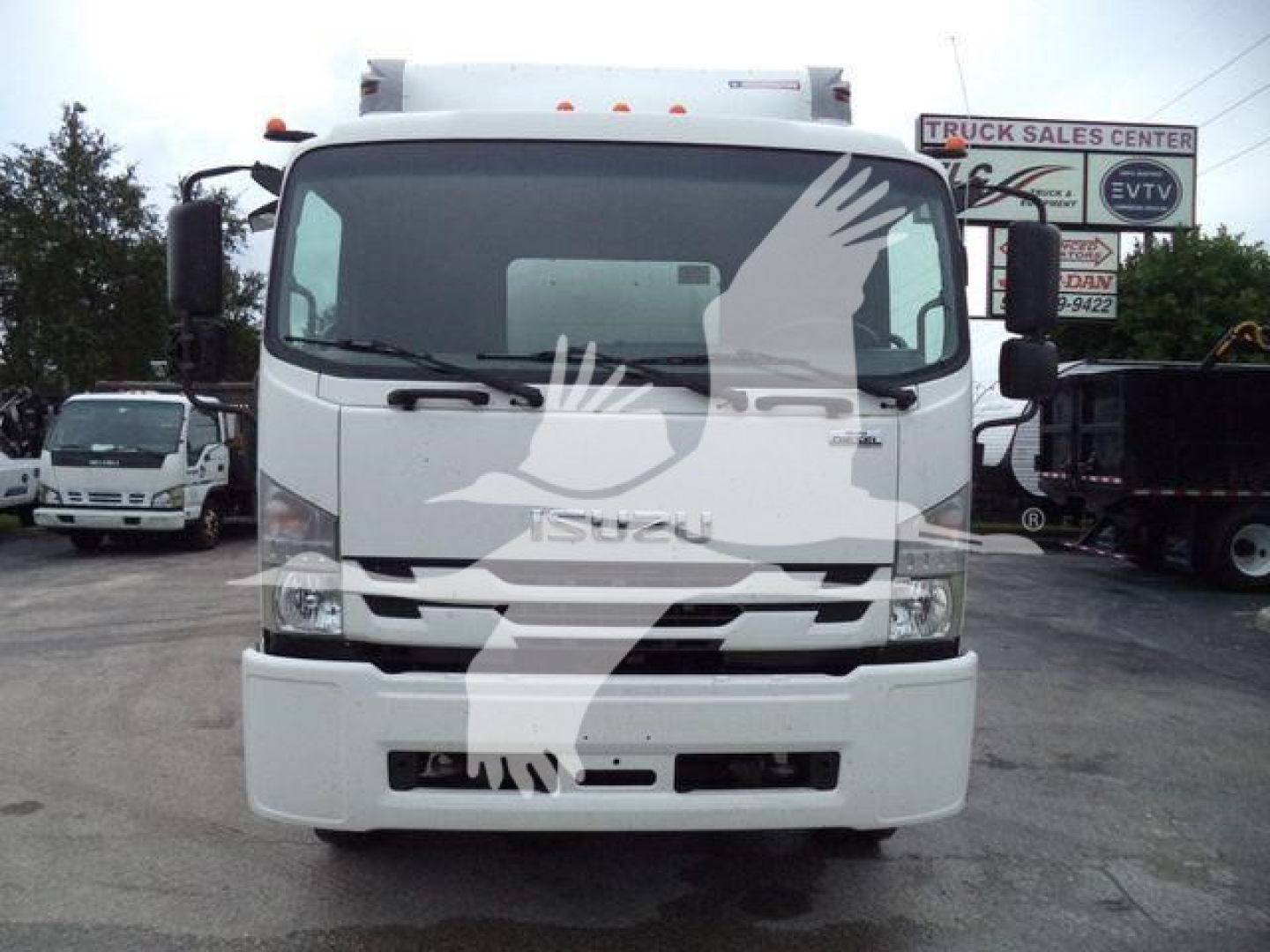 Isuzu truck front view with windshield wipers, grille, and bumper. The image shows a white Isuzu medium-duty box truck with stock number and dealership signage in the background.