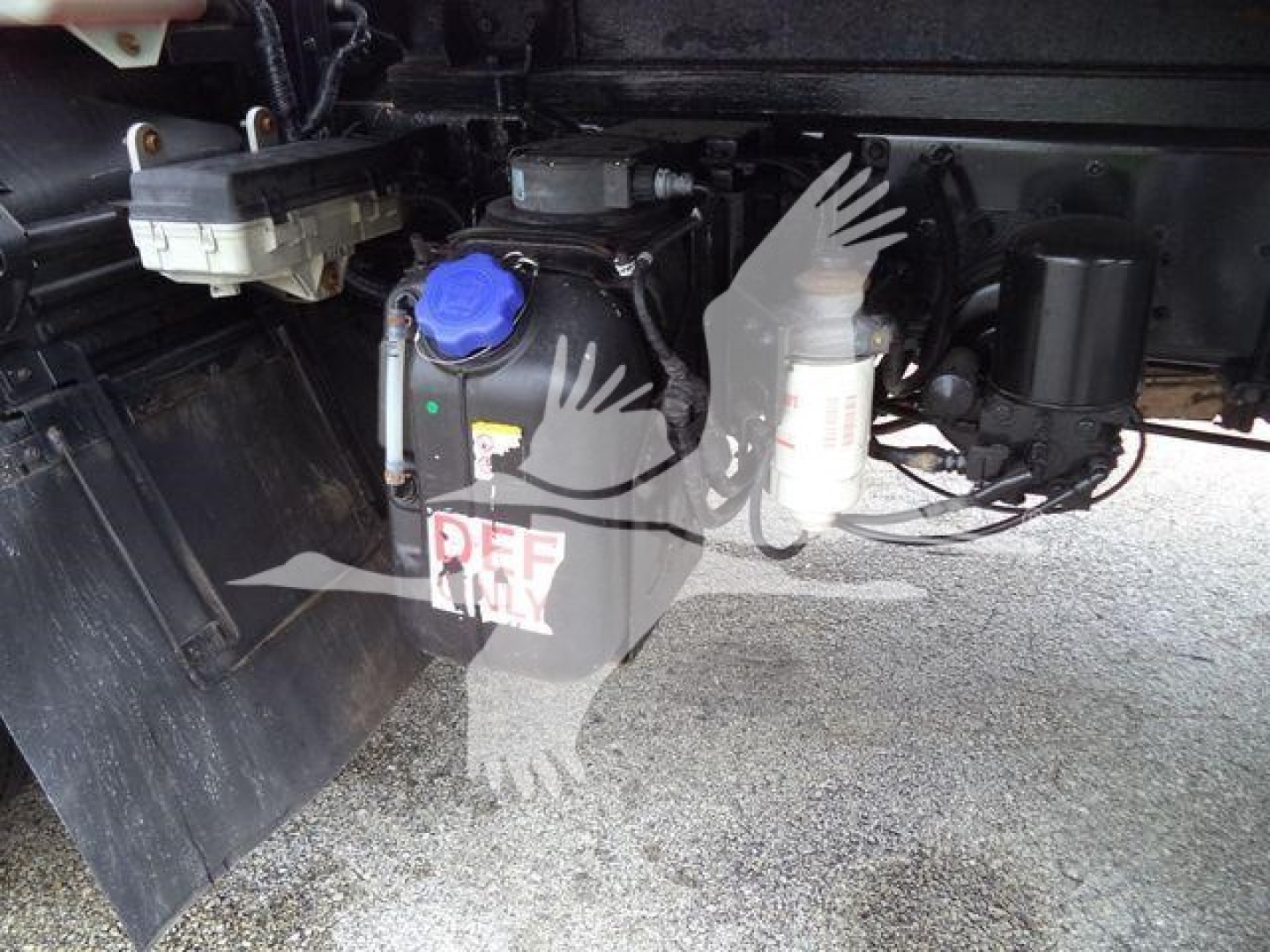 Isuzu truck undercarriage showing coolant reservoir with blue cap and DEF container, hoses and wiring visible.