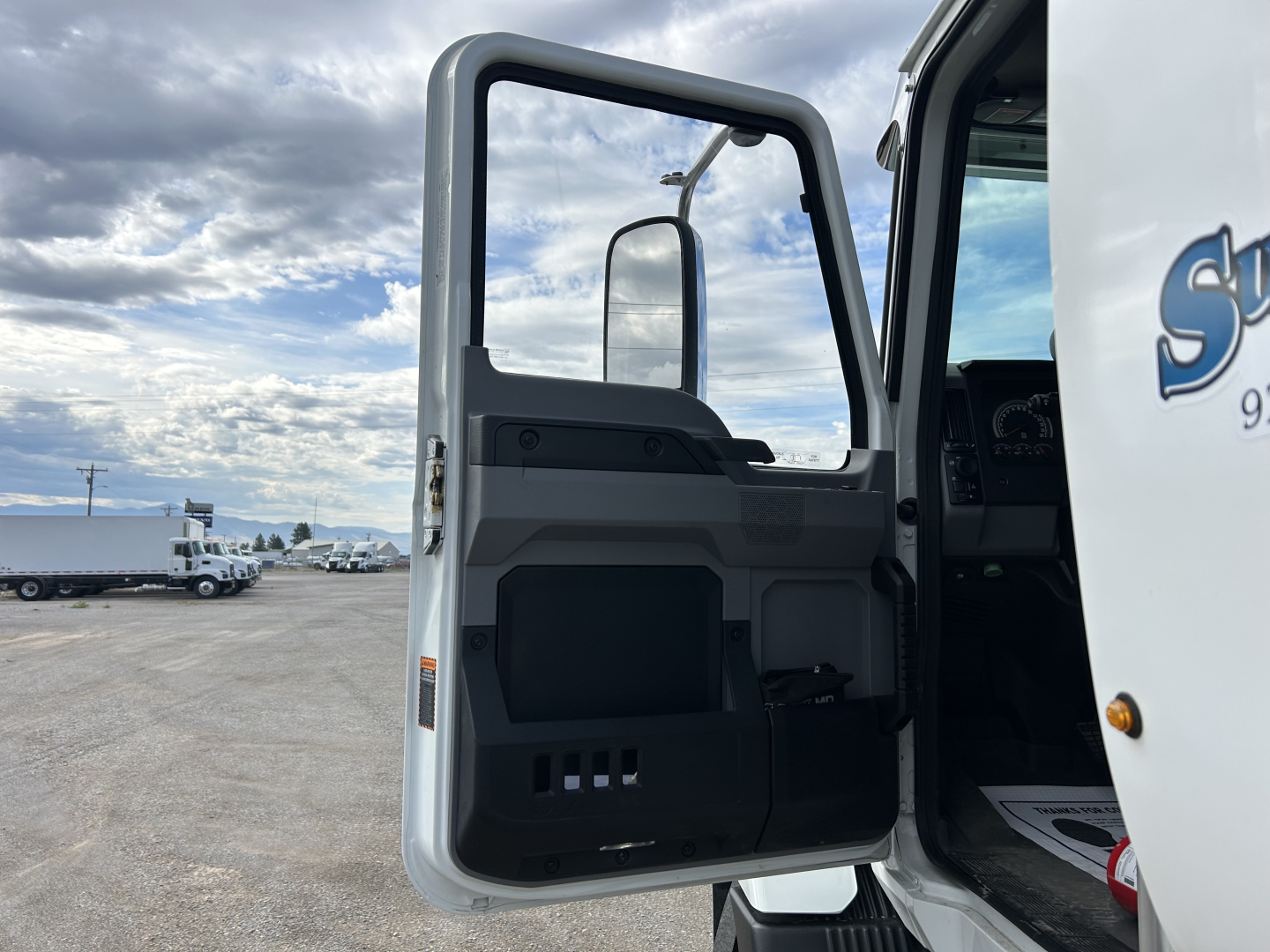 Truck cab interior door view of a 2025 Mack MD7 with open door, showing interior panel, window, and steps; part of the exterior white cab with blue graphics visible.