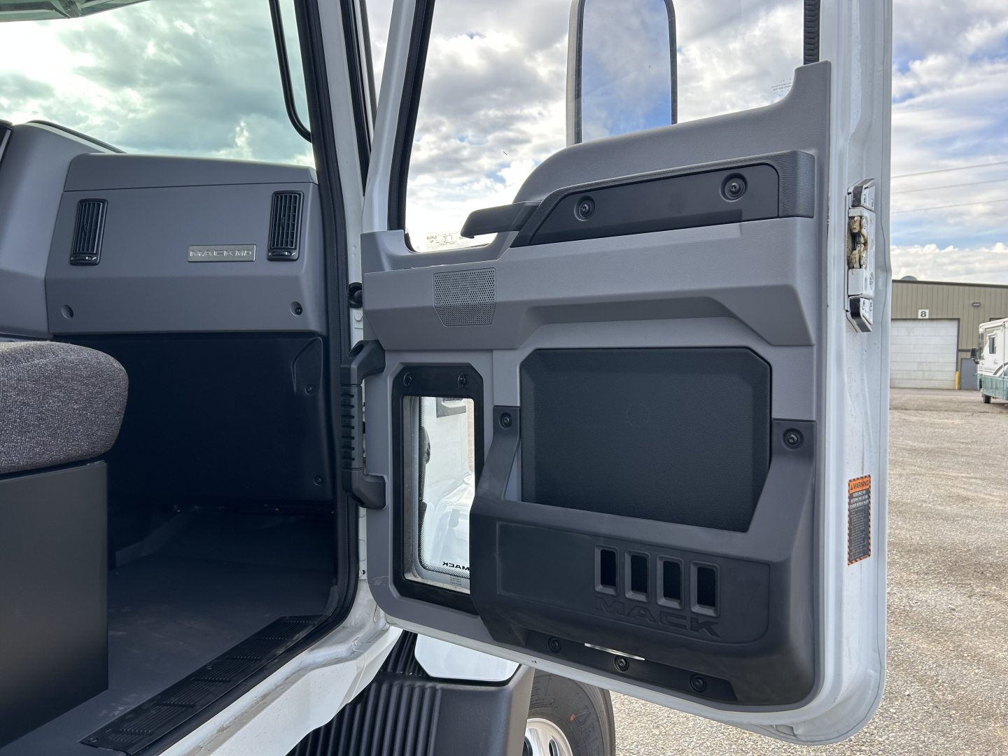 Truck cab interior door panel of a 2025 Mack MD7 showing storage pockets and control panel, with window and exterior environment in background.