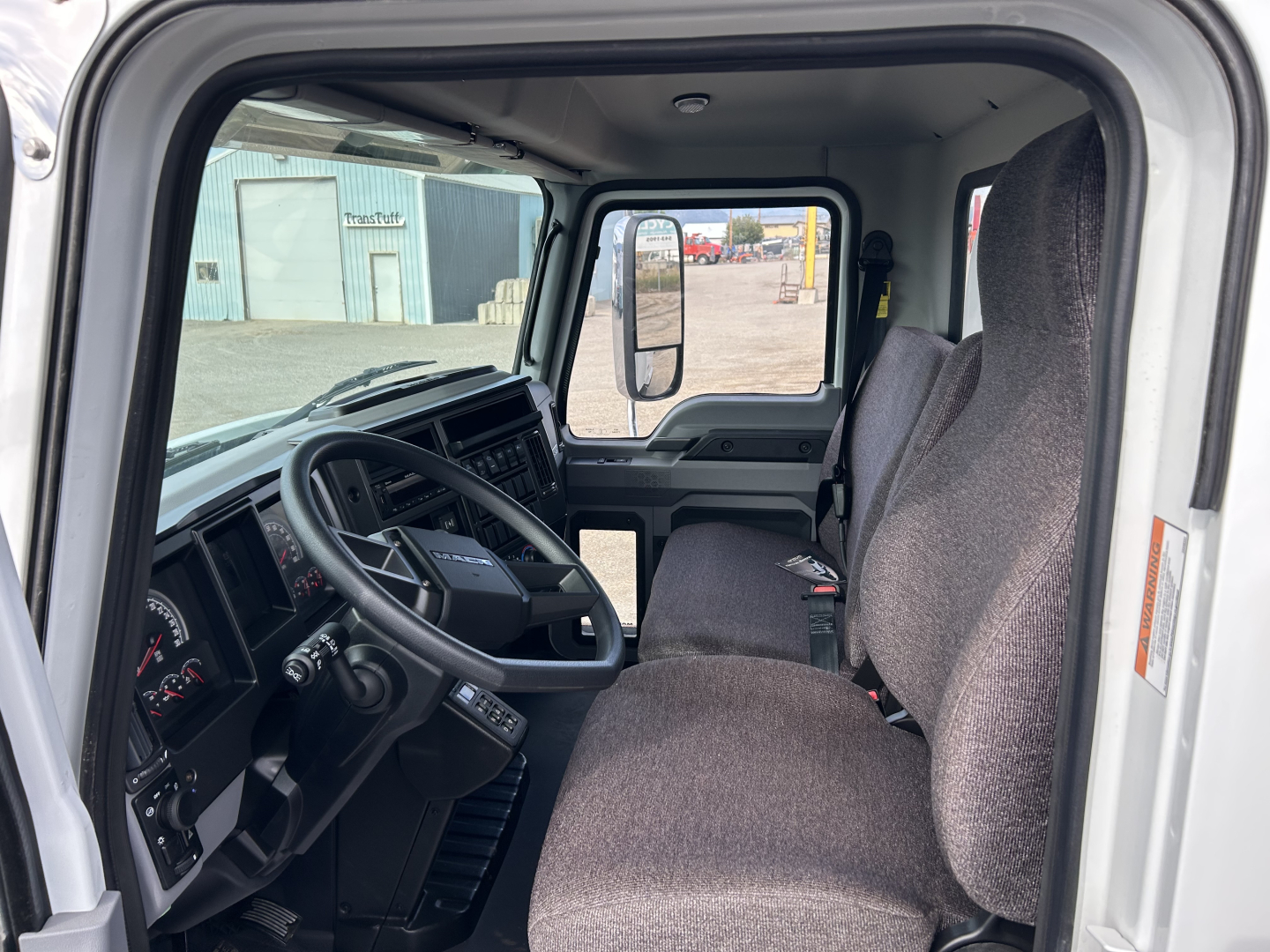 Truck cab interior of a 2025 Mack MD7 showing driver seat and passenger seat, steering wheel, dashboard, and side mirror visible from the open door.
