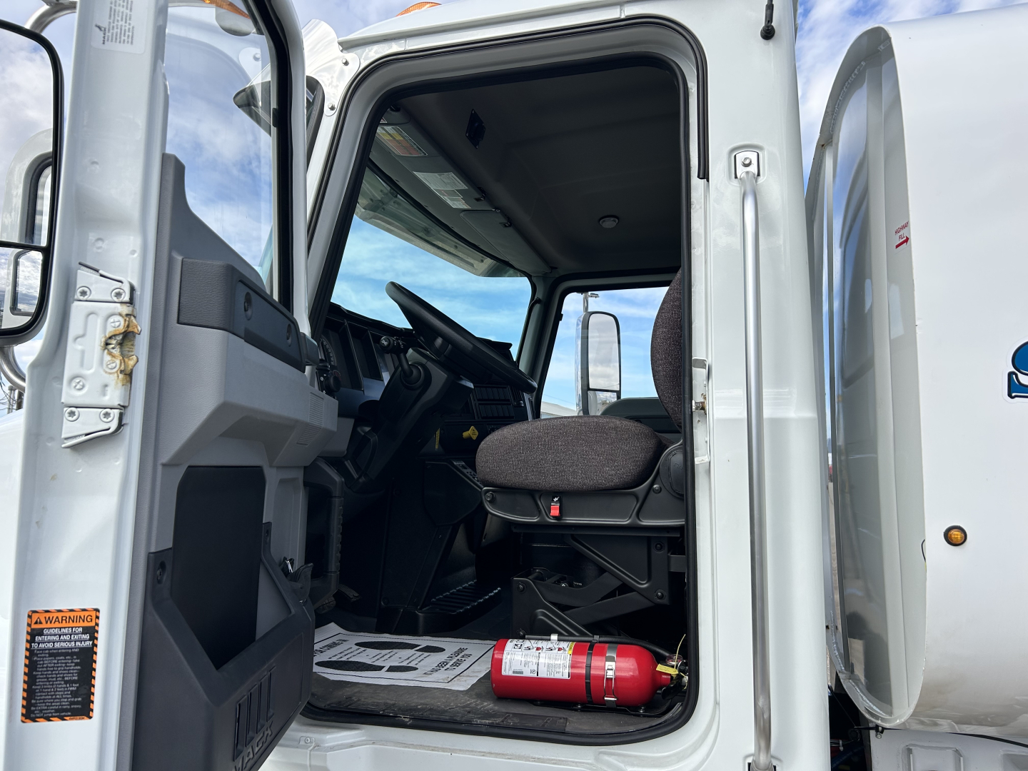 Truck cab interior of a 2025 Mack MD7 shown from the open driver door, with steering wheel, dashboard, seat, and a fire extinguisher on the floor.