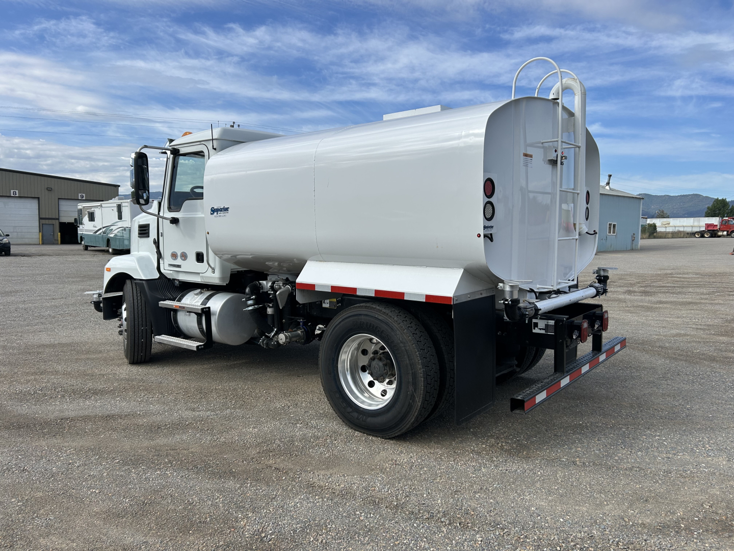 Side view of a white 2025 Mack MD7 water tanker truck with cylindrical tank, rear access ladder, and dual rear wheels, showing brand text on cab and rear bumper.