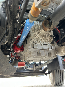 Transmission/drive area of a 2025 Mack MD7 showing the transfer case, blue hydraulic line protector, and surrounding hoses and components.