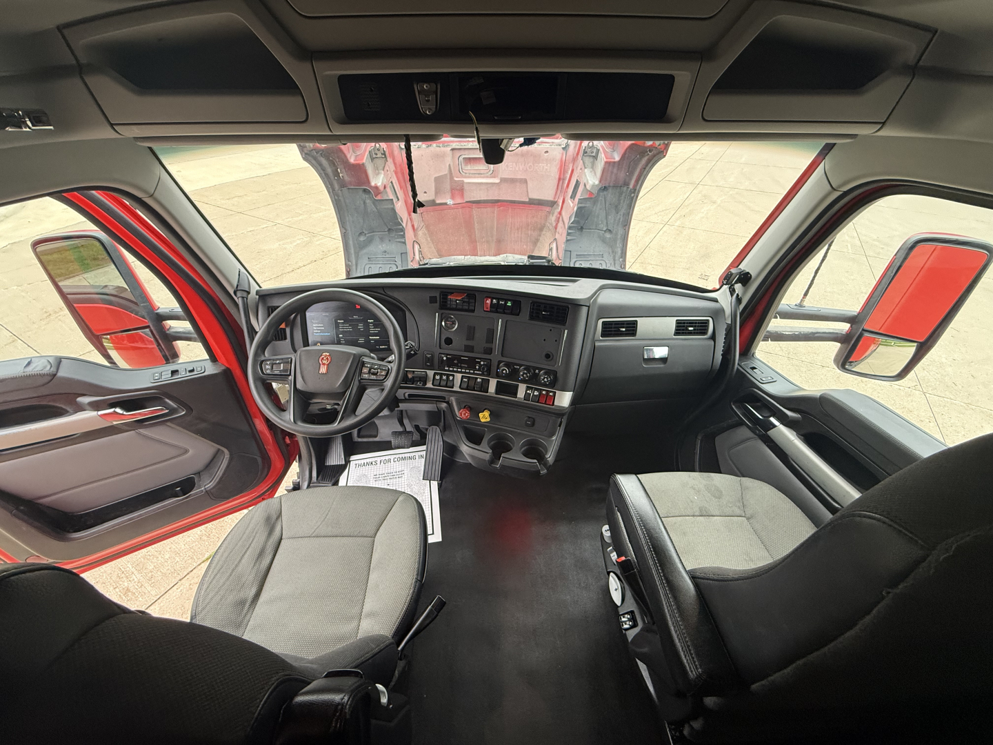 Kenworth T680 cab interior showing steering wheel, dashboard with multiple gauges and controls, driver and passenger seats, center console.