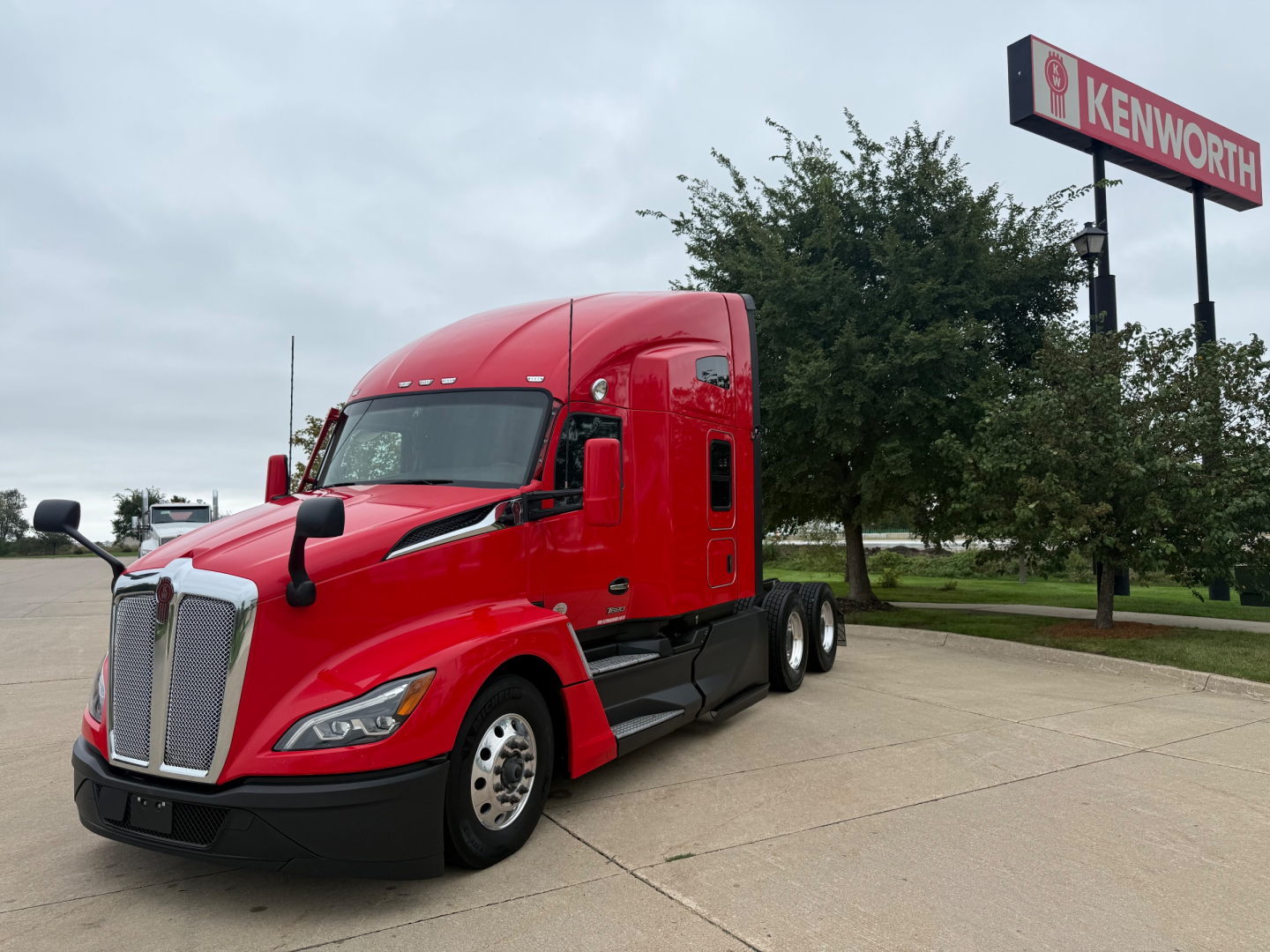 Kenworth T680 conventional sleeper truck in red parked in lot showing front and side with Kenworth sign in background.