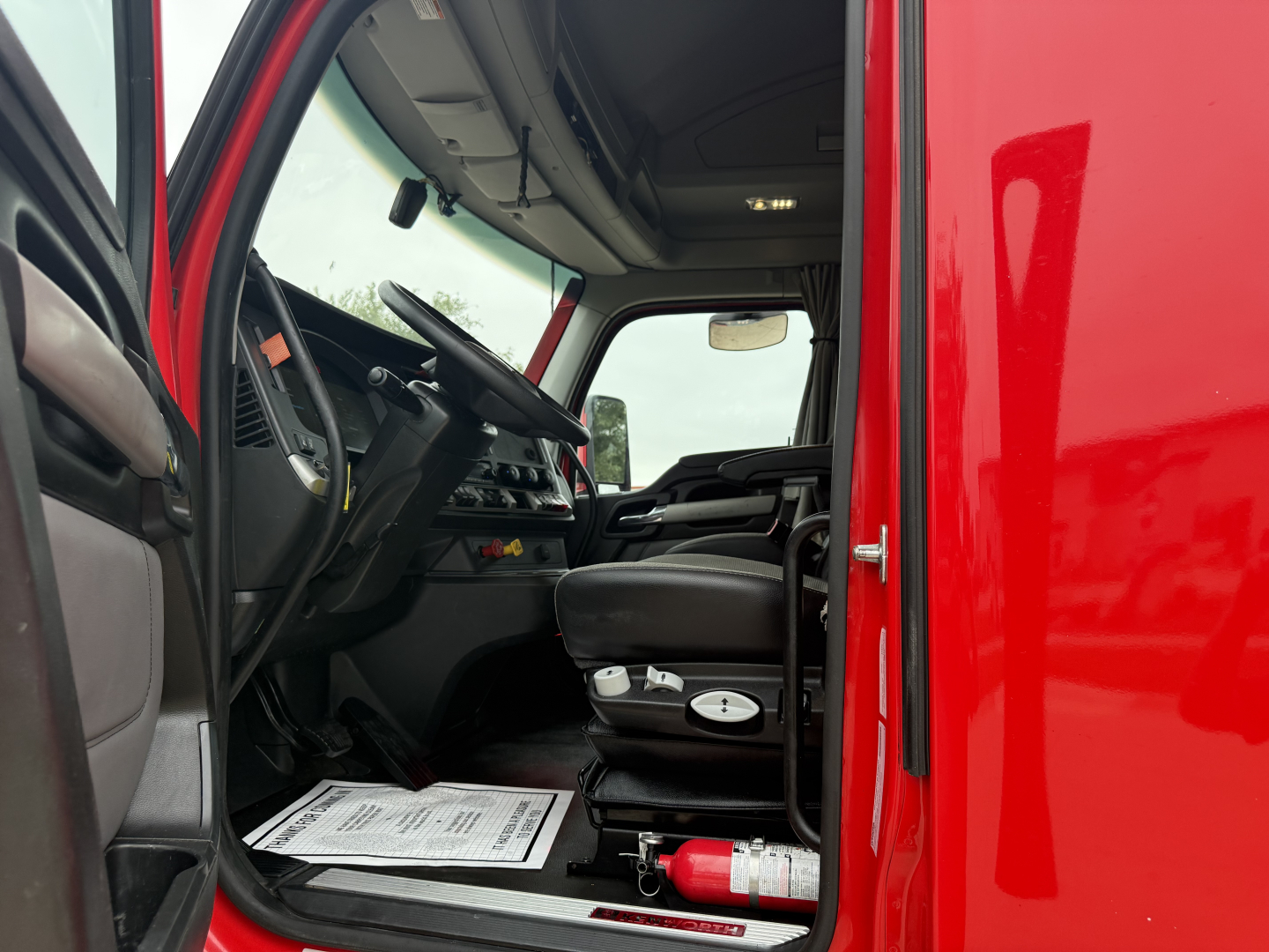 Kenworth T680 truck interior cab view from open driver door showing steering wheel and dashboard.