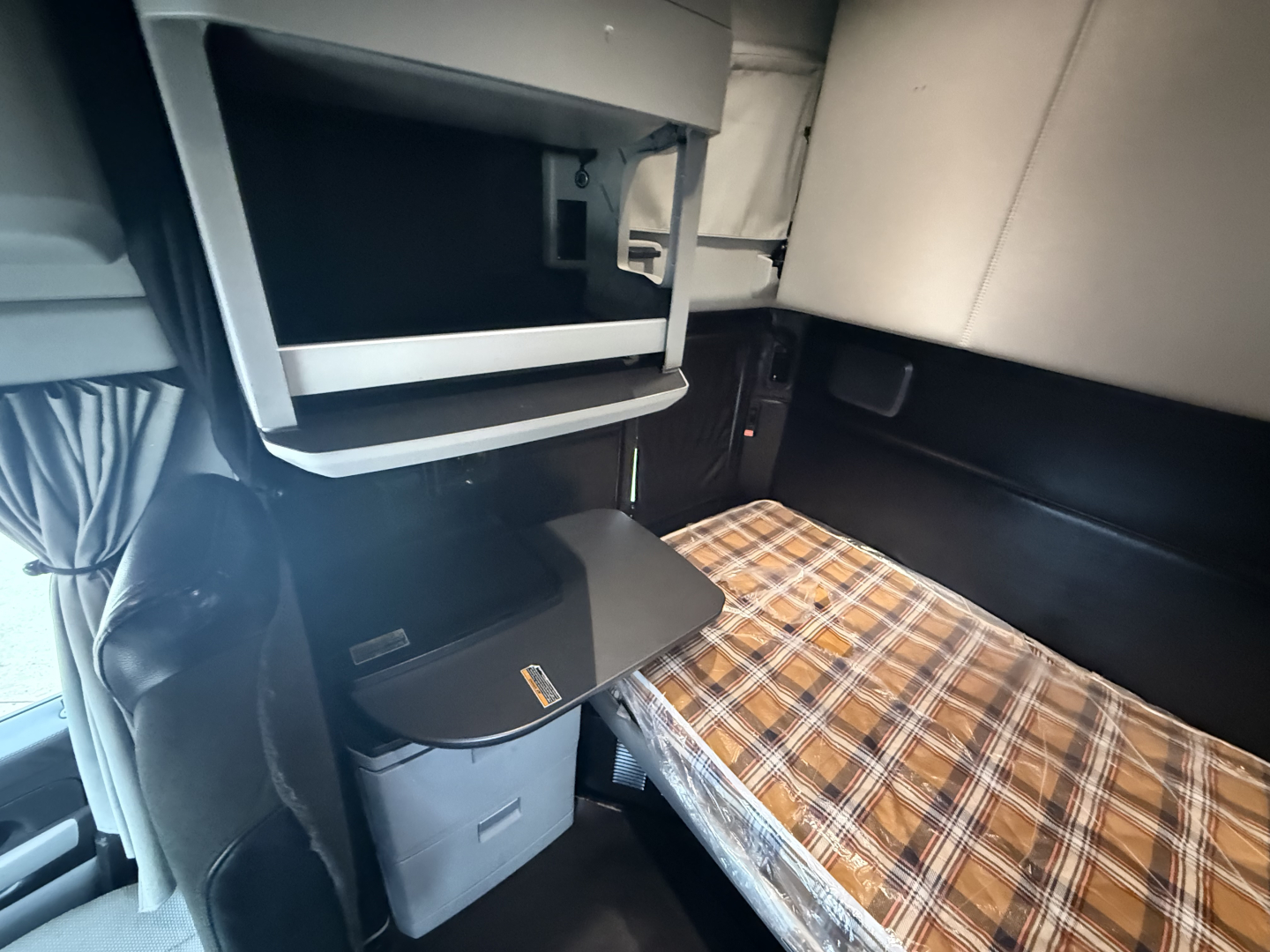 Truck cab interior with sleeper area and plaid mattress cover; overhead compartments and a small cabinet/locker, plastic storage bin, and a pull-out tray.