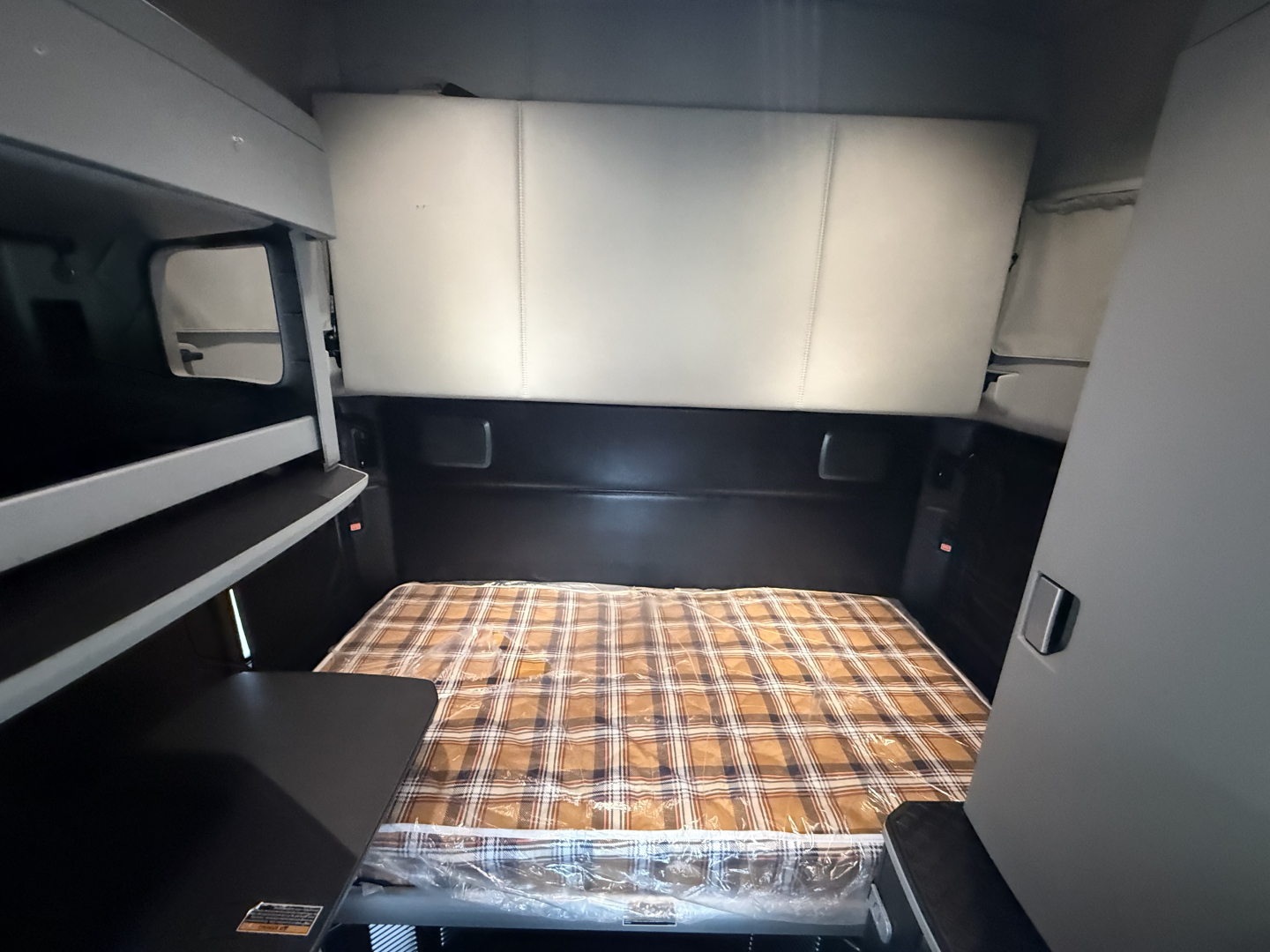 Kenworth T680 sleeper cab interior showing bunk bed with plaid mattress and side storage compartments.