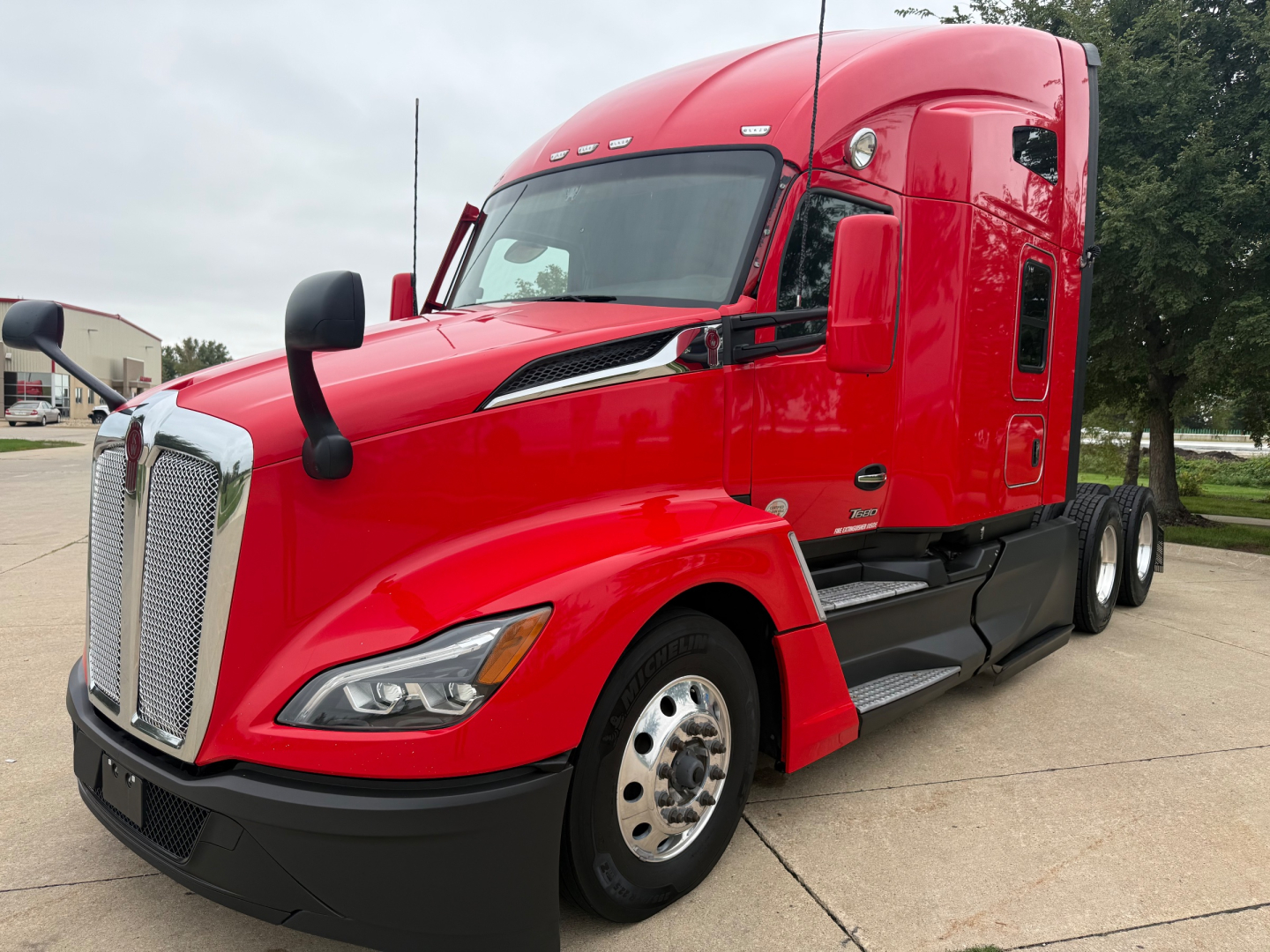 Kenworth T680 conventional sleeper truck showing red cab and front grille with black bumper and dual rear wheels.
