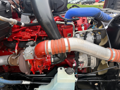Engine bay of a 2023 Kenworth T680 showing red engine block, large intake pipe, turbocharger, silver exhaust pipe, blue silicone hose, and coolant reservoir.