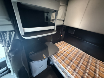 Truck cab interior with sleeper area and plaid mattress cover; overhead compartments and a small cabinet/locker, plastic storage bin, and a pull-out tray.
