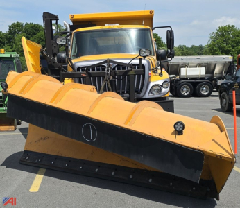 Dump truck with front-mounted snow plow blade in a dealership lot, yellow vehicle with black grille and plow in foreground.