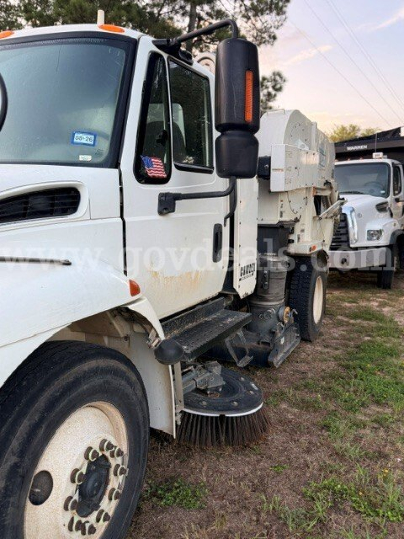 International 4300 street sweeper dump truck side view showing front wheel, body, and sweeping equipment. Visible text includes a small USA flag sticker on the door and generic vehicle labels.