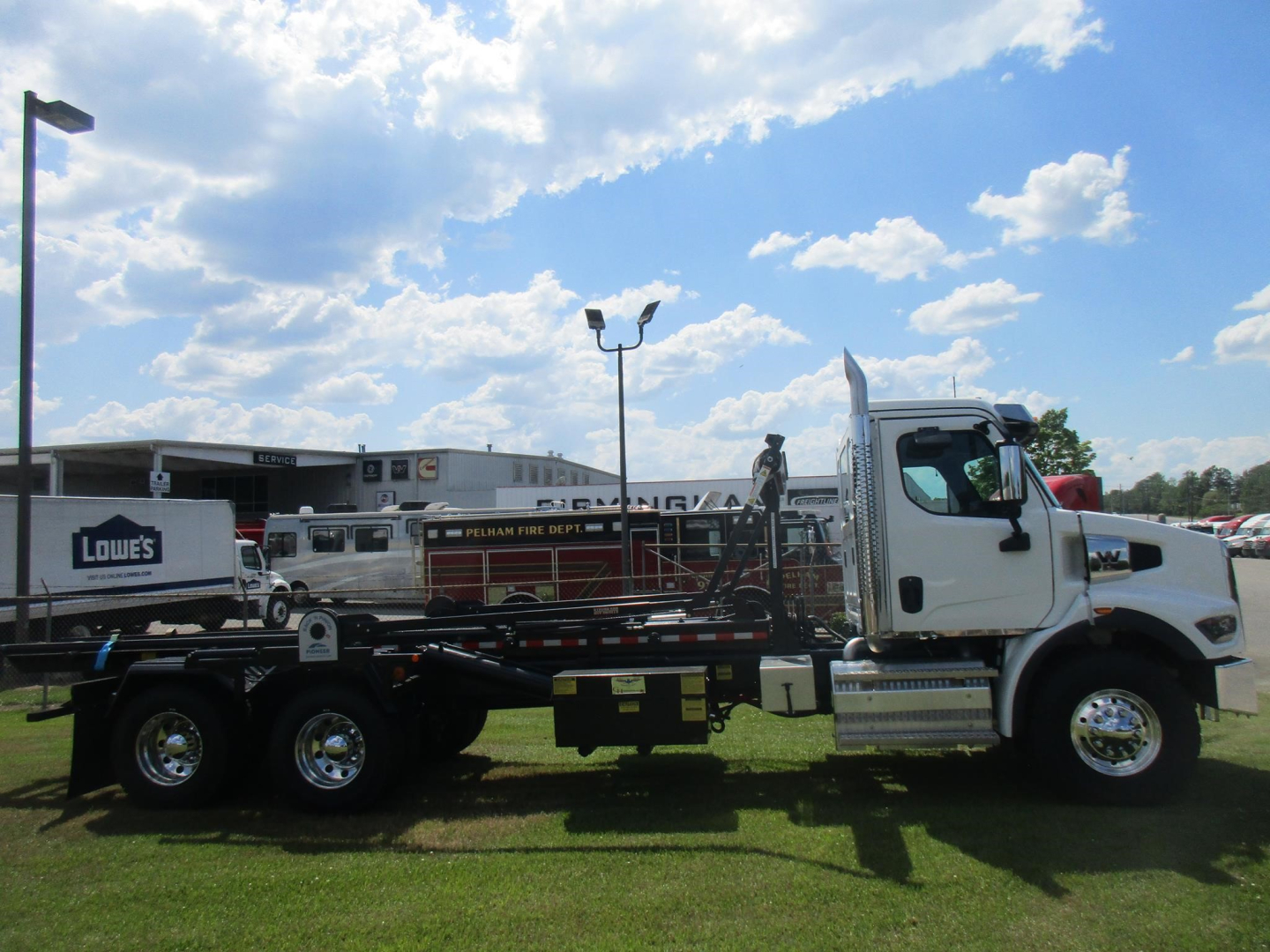 2026 Western Star 47X roll-off truck side view showing cab and flatbed with equipment and trailer wheels, blue sky with clouds.