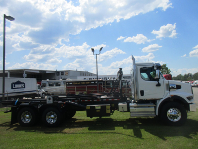 2026 Western Star 47X roll-off truck side view showing cab and flatbed with equipment and trailer wheels, blue sky with clouds.