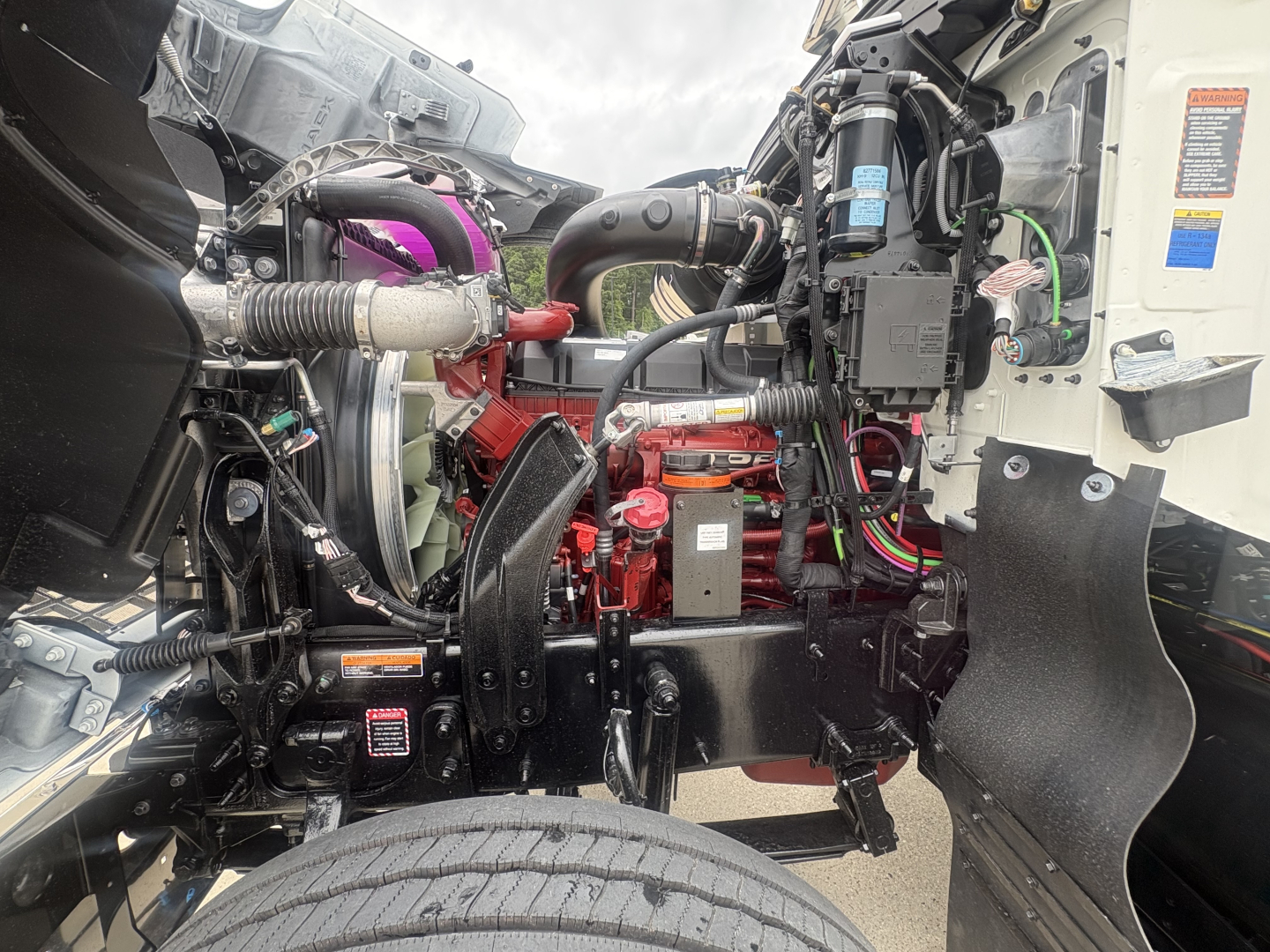 Truck engine bay of 2027 Mack PI64T showing red engine block, black chassis, various hoses, wiring, and a pink coolant hose visible.