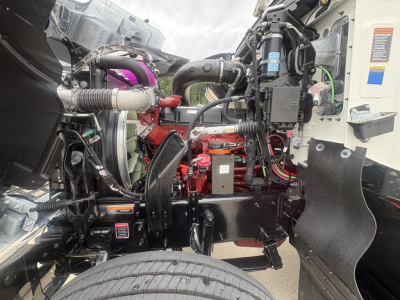 Truck engine bay of 2027 Mack PI64T showing red engine block, black chassis, various hoses, wiring, and a pink coolant hose visible.